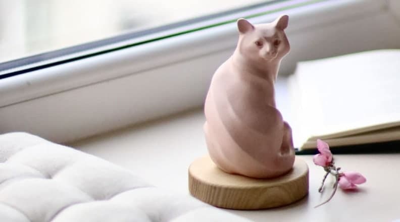 A pink remembrance stone shaped as a seated cat, on a round wooden base by a window